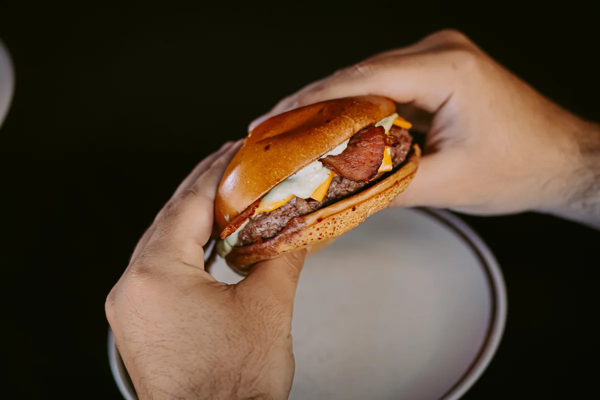 Hambúrguer sendo segurado com as duas mãos, com bacon, cheddar e maionese verde